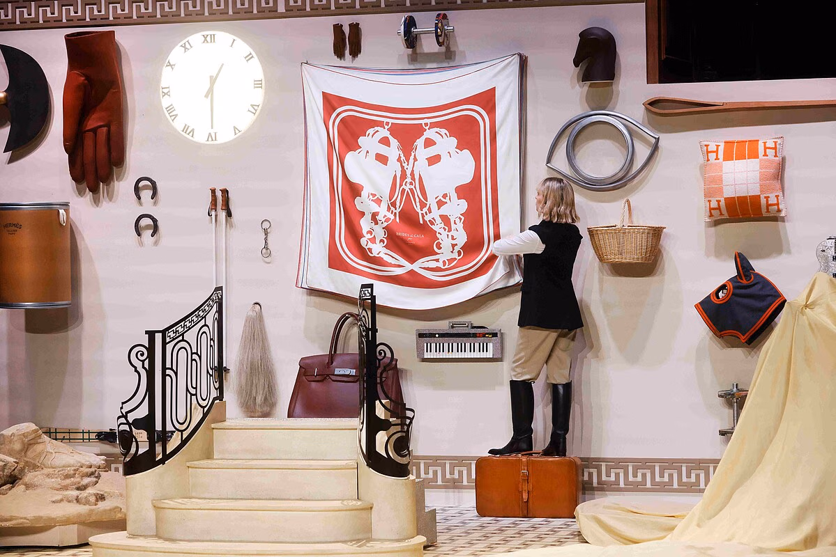 A performer on a suitcase before a wall of scarves and objects