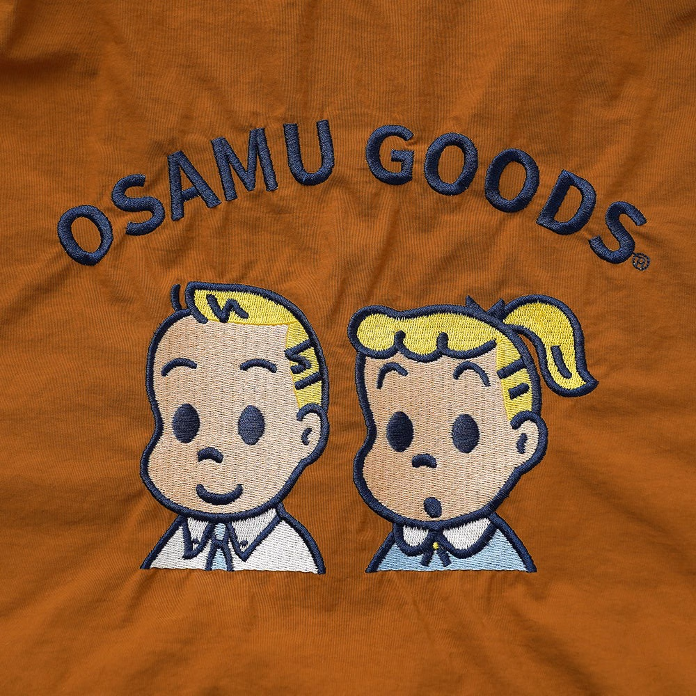 ภาพใกล้ลายปักแจ็กกับจิลบนผ้าคาเมลพร้อมข้อความ OSAMU GOODS เดิม